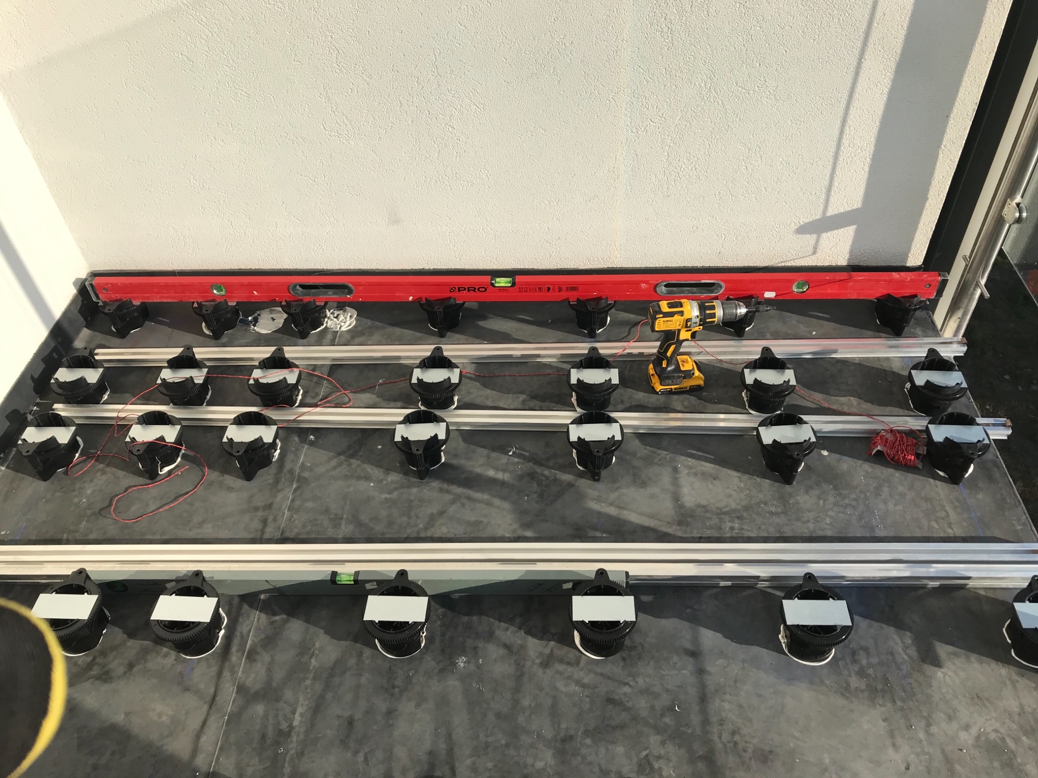 Przygotowanie do montażu podłogi: poziomica PRO, wkrętarka DeWalt i elementy konstrukcyjne z regulowaną wysokością na szarym podłożu przy białej ścianie.