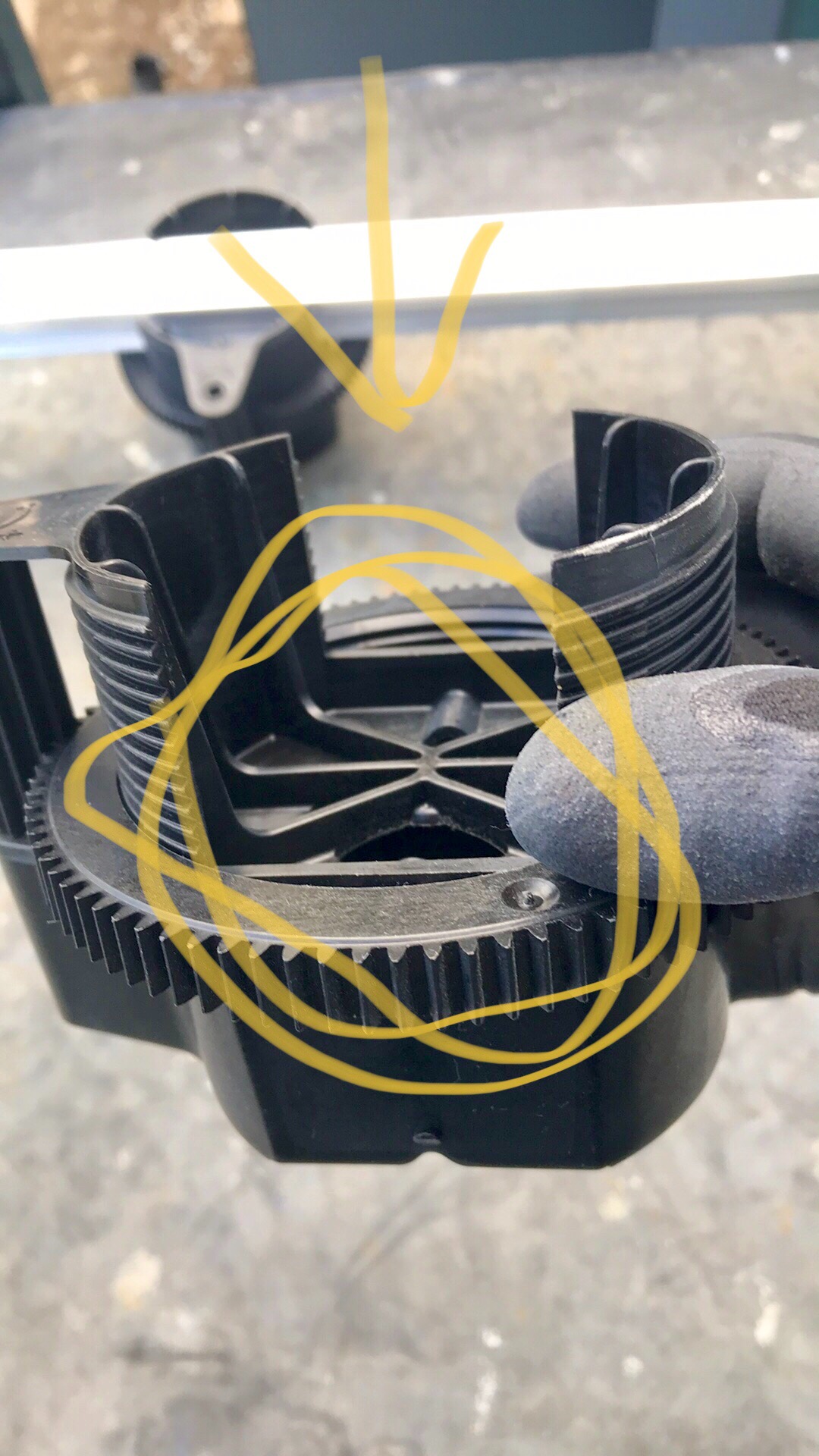 Czarny element mocujący z zębatym obwodem i fragmentami gumy, prawdopodobnie do regulacji wysokości, na tle szarej powierzchni.