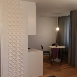 I.O.EXPERT Sp.zo.o - Nowoczesne wnętrze mieszkania z dekoracyjną ścianą z okrągłych elementów, widok na jadalnię z okrągłym stołem i szarymi zasłonami.