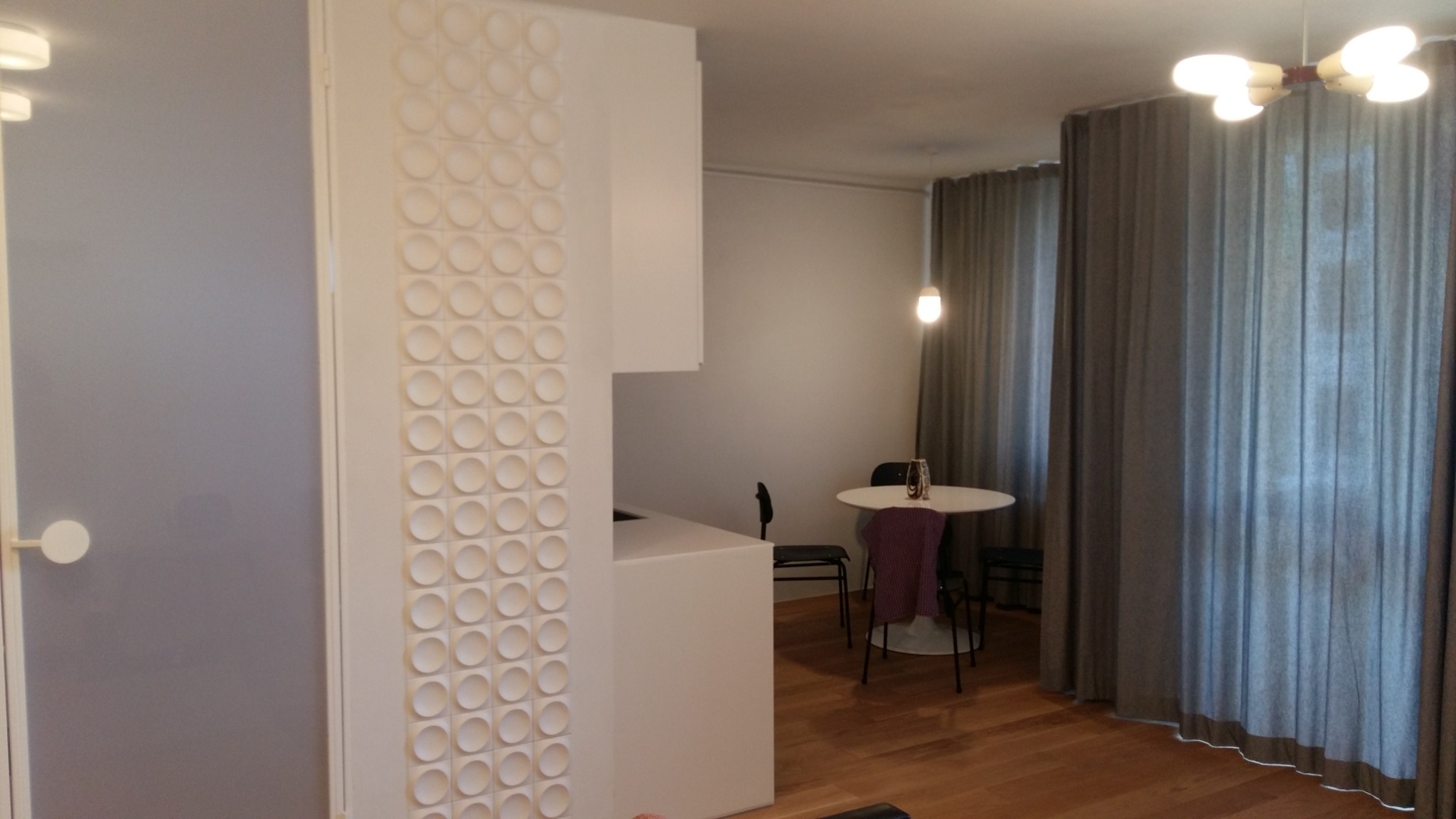 Nowoczesne wnętrze mieszkania z dekoracyjną ścianą z okrągłych elementów, widok na jadalnię z okrągłym stołem i szarymi zasłonami.