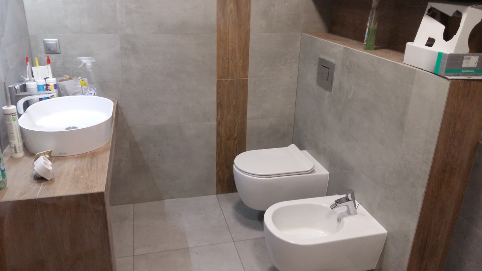 Nowoczesna łazienka z szarą płytką, drewnianym akcentem, wiszącą miską WC i bidetem oraz umywalką nablatową. Na blacie widoczne akcesoria łazienkowe i materiały montażowe.