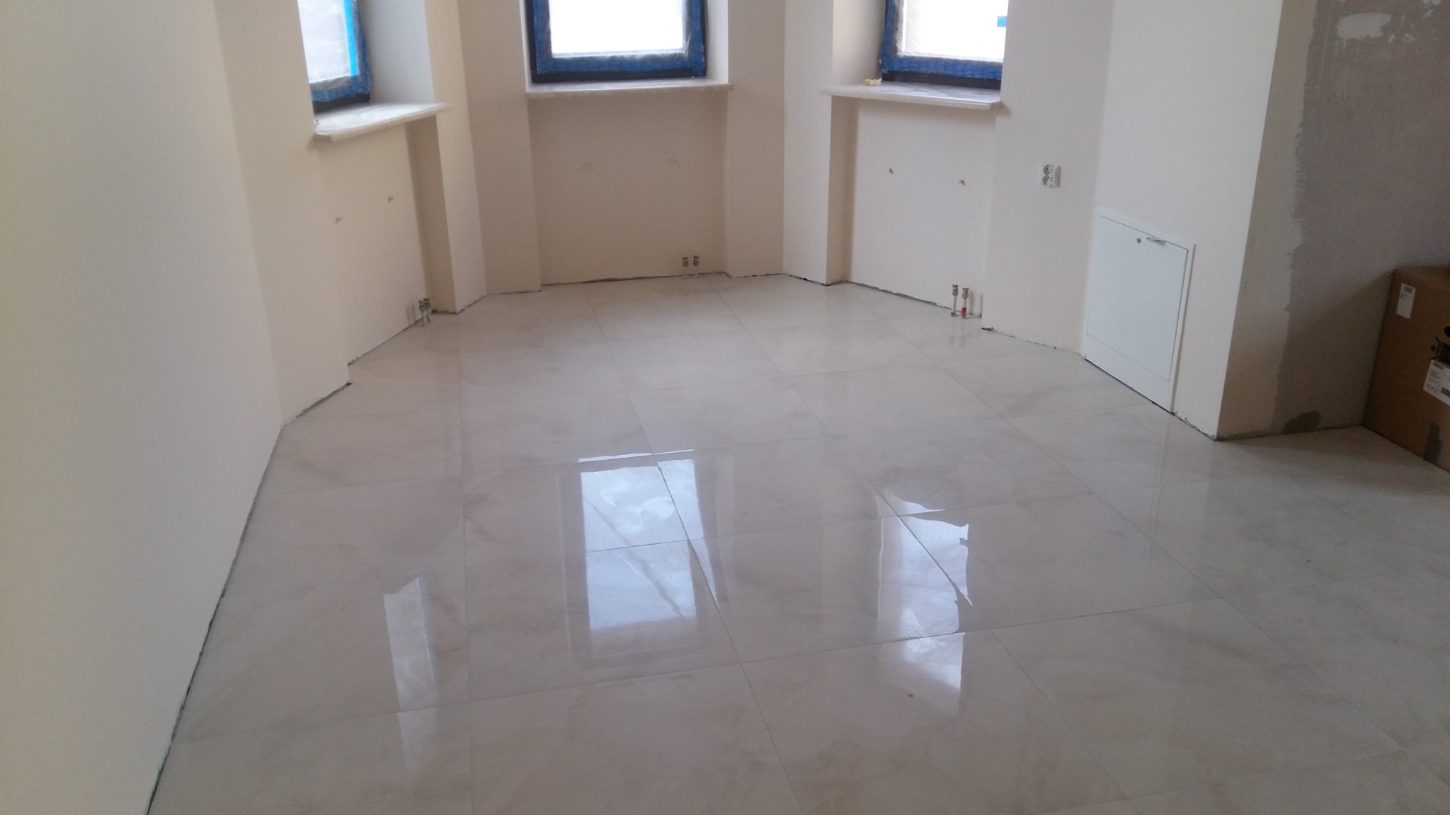 Wykończone wnętrze z jasnymi, błyszczącymi płytkami podłogowymi, świeżo pomalowanymi ścianami w odcieniu kremowym, widocznymi instalacjami pod oknami i kartonem w rogu pomieszczenia.