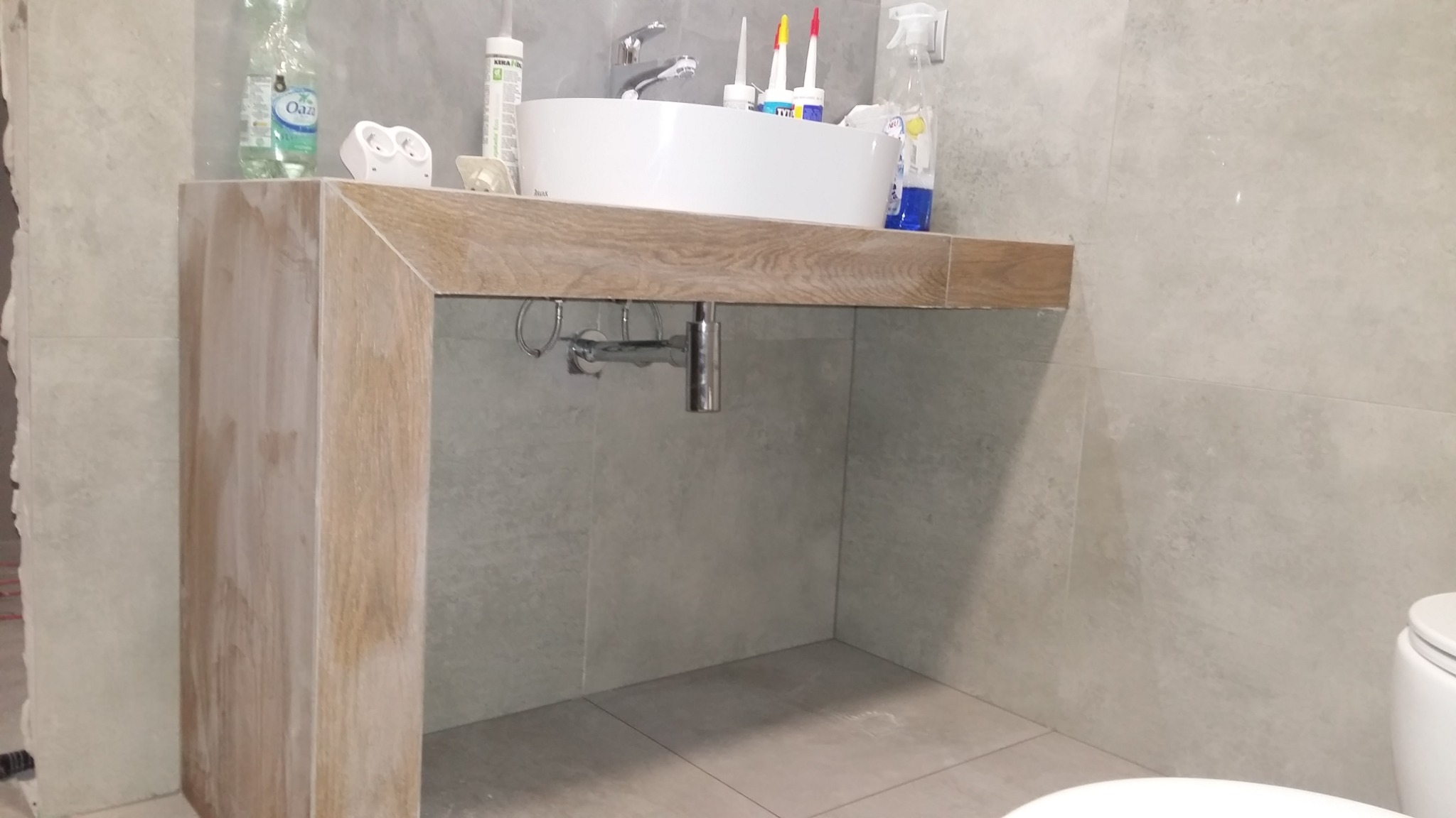 Nowoczesna umywalka nablatowa z drewnianym blatem i szafką w łazience wykończonej szarymi płytkami, widoczne akcesoria montażowe i chemia budowlana.