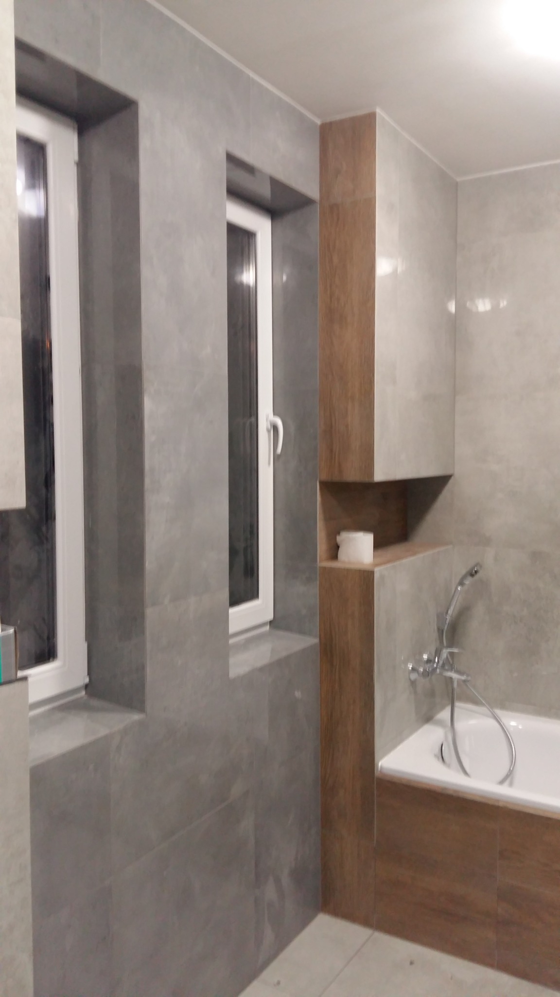 Wykończona łazienka z szarymi płytkami imitującymi kamień, zabudową w kolorze drewna, białą wanną i oknami z białymi ramami.