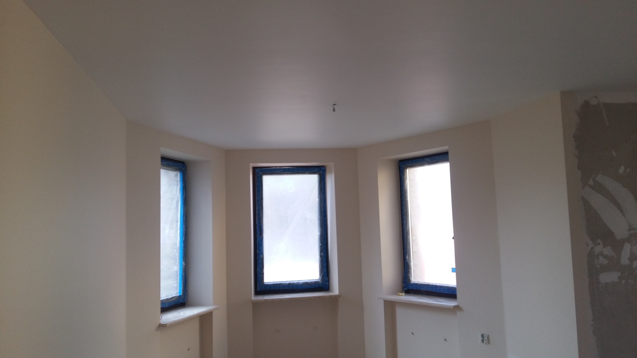 Pokój po świeżym malowaniu na jasny odcień, z trzema oknami zabezpieczonymi folią malarską, na ścianie widoczne ślady po wcześniejszych pracach wykończeniowych.