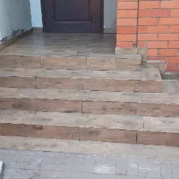 Schody wejściowe wyłożone płytkami imitującymi drewno, z widocznymi niedoskonałościami przy krawędziach i ścianie, obok czerwona ławka z wiadrem i mopem.