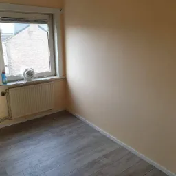 Pomalowane na beżowo wnętrze pokoju z szarą podłogą z paneli, białym oknem, grzejnikiem i wentylatorem na parapecie.