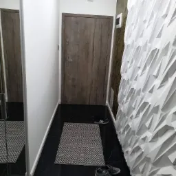 Nowoczesny przedpokój z czarną marmurową podłogą, drewnianymi drzwiami i białą ścianą dekoracyjną o geometrycznym wzorze 3D, uzupełniony o dywanik i parę butów.