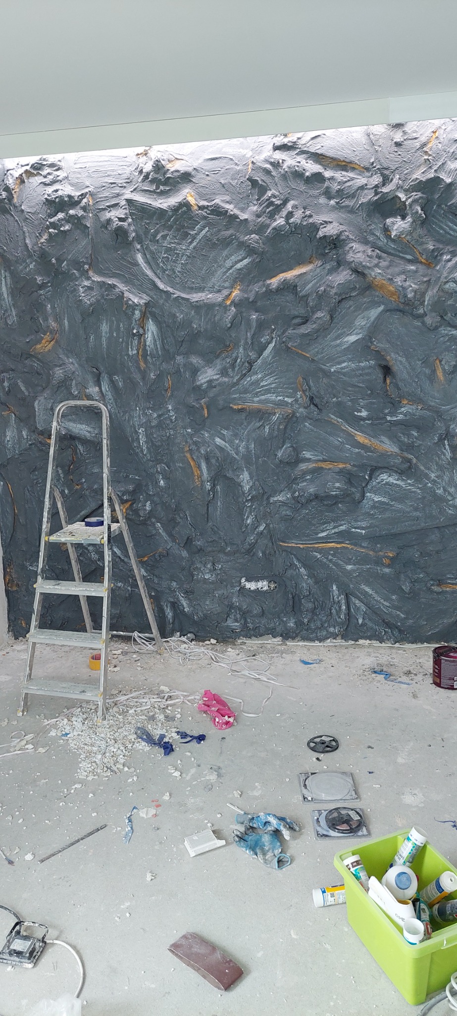 Szara ściana z fakturą imitującą kamień, miejscami z akcentami złota, widoczna drabina malarska i akcesoria malarskie na podłodze w trakcie wykonywania prac.