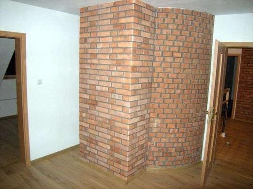 Wnętrze z dekoracyjną ścianą z czerwonej cegły, zaokrąglony narożnik, drewniana podłoga i ościeżnice drzwiowe.