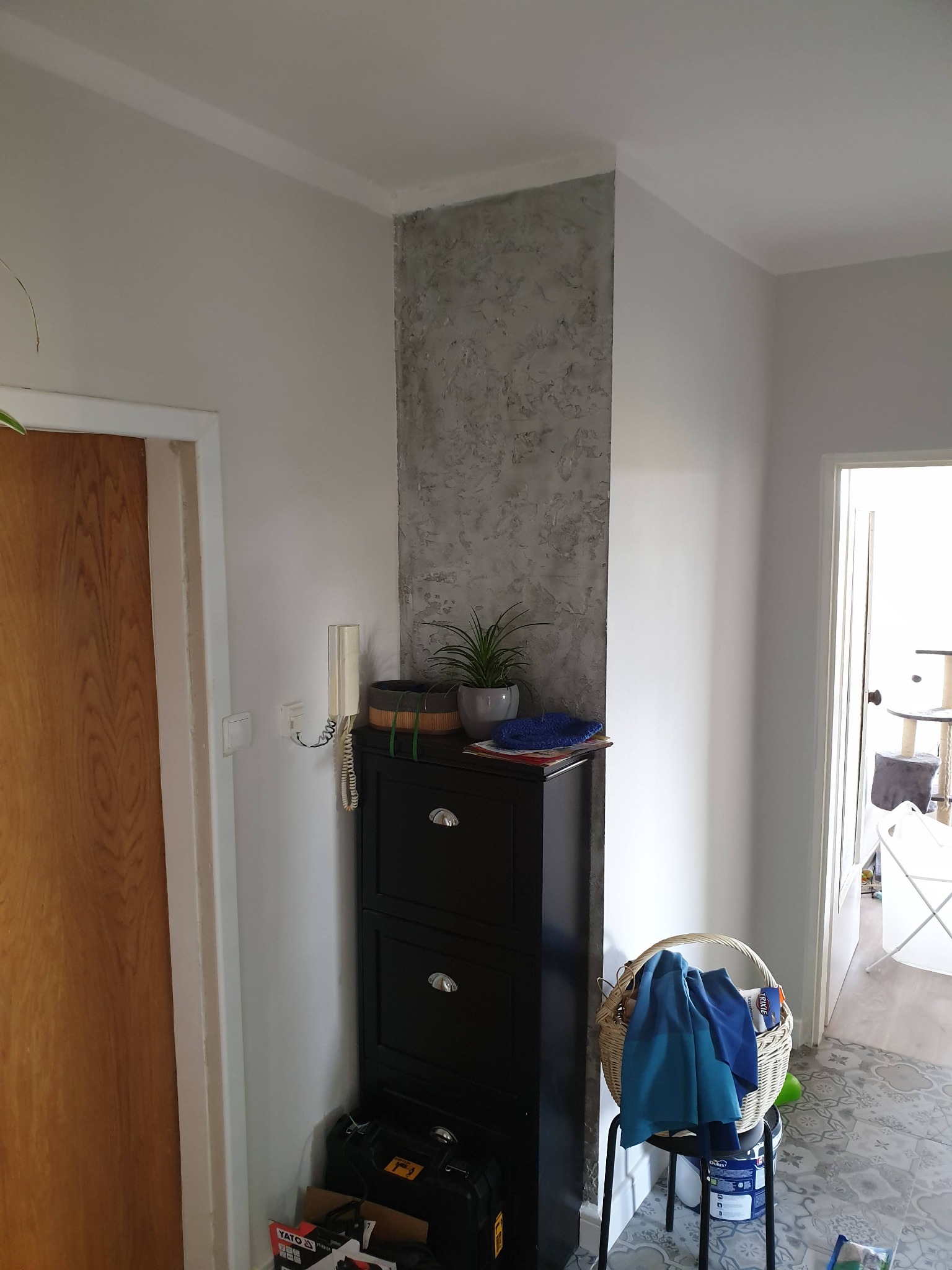 Fragment korytarza z dekoracyjną ścianą w odcieniach szarości, kontrastującą z gładką, jasną ścianą; widoczna szafka na buty, roślina doniczkowa i kosz z niebieskimi tkaninami.