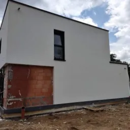 Dwukondygnacyjny budynek z fragmentem elewacji w trakcie prac: otynkowana, biała ściana kontrastuje z surowym murem z czerwonej cegły, widoczne okno z czarną ramą, fundamenty i niezagospodarowany...