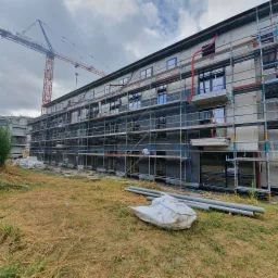 Wielopiętrowy budynek w trakcie ocieplania elewacji, widoczne rusztowania, warstwa izolacyjna i rury wentylacyjne na ścianie, żuraw budowlany w tle.