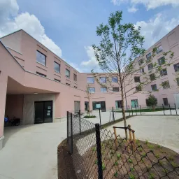 Nowoczesny budynek o różowej elewacji widziany z dziedzińca z młodymi drzewami, czarnym ogrodzeniem i błękitnym niebem z chmurami.