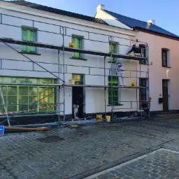 Dwóch pracowników na rusztowaniu maluje elewację budynku. Okna zabezpieczone folią, widoczne wiadra z farbą marki STO.