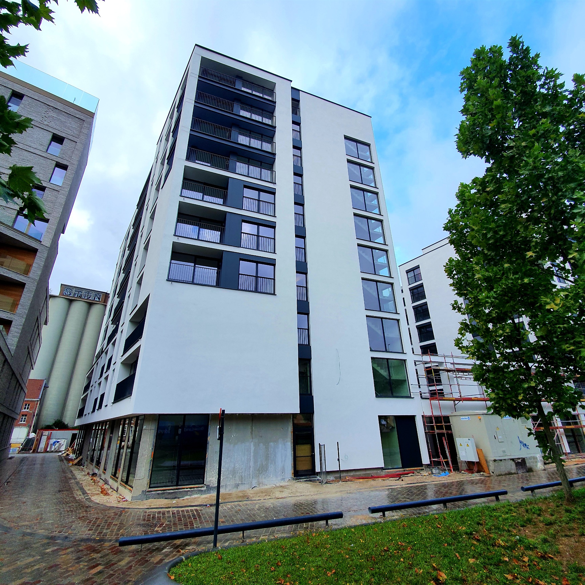 Wysoki, nowo wybudowany budynek mieszkalny z białymi ścianami i czarnymi balkonami, widziany z dołu, z elementami rusztowania i prac wykończeniowych w dolnej części elewacji.