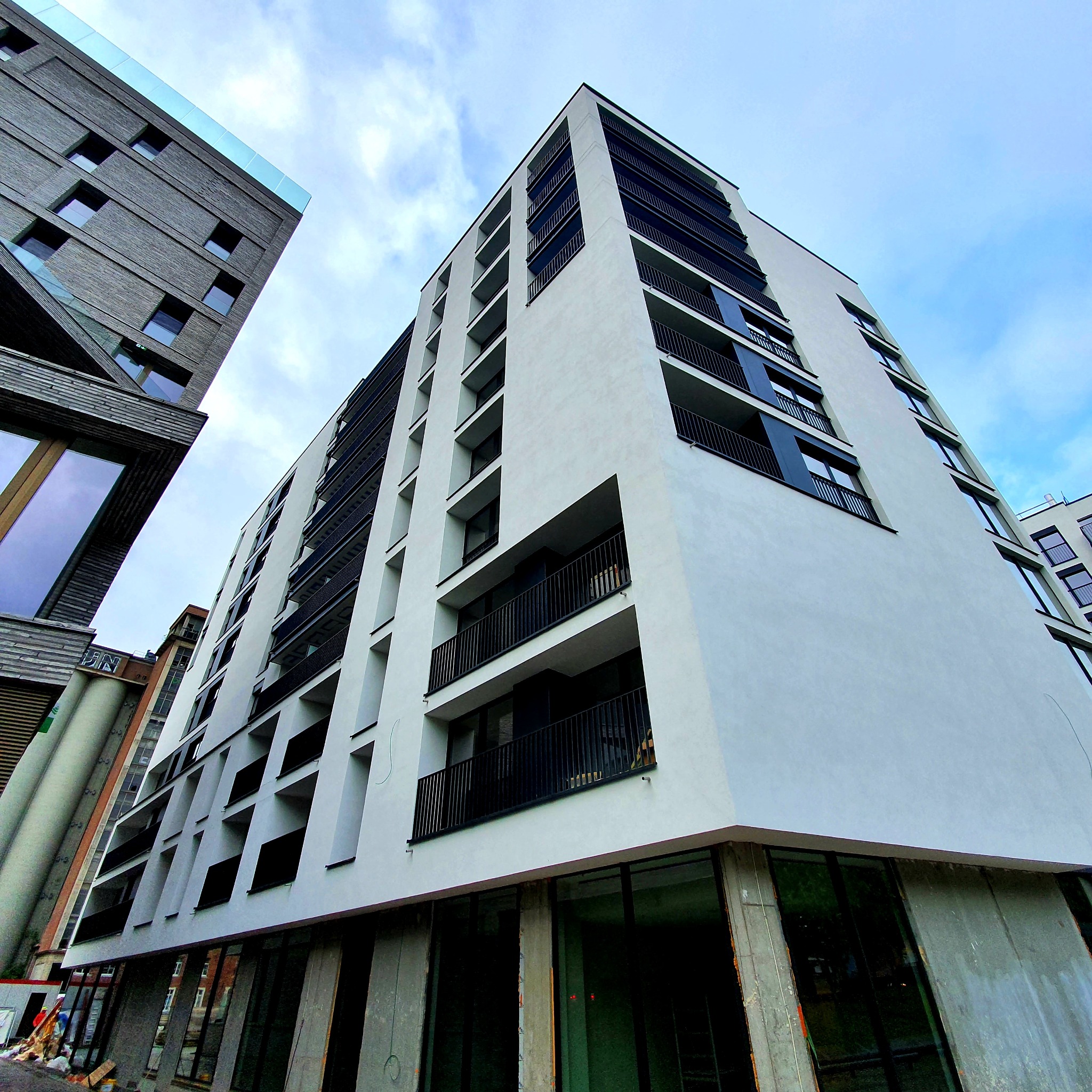 Nowoczesny budynek wielorodzinny z białymi ścianami i czarnymi balkonami, widok z dołu na tle błękitnego nieba, w dolnej części widoczne surowe wykończenia parteru.