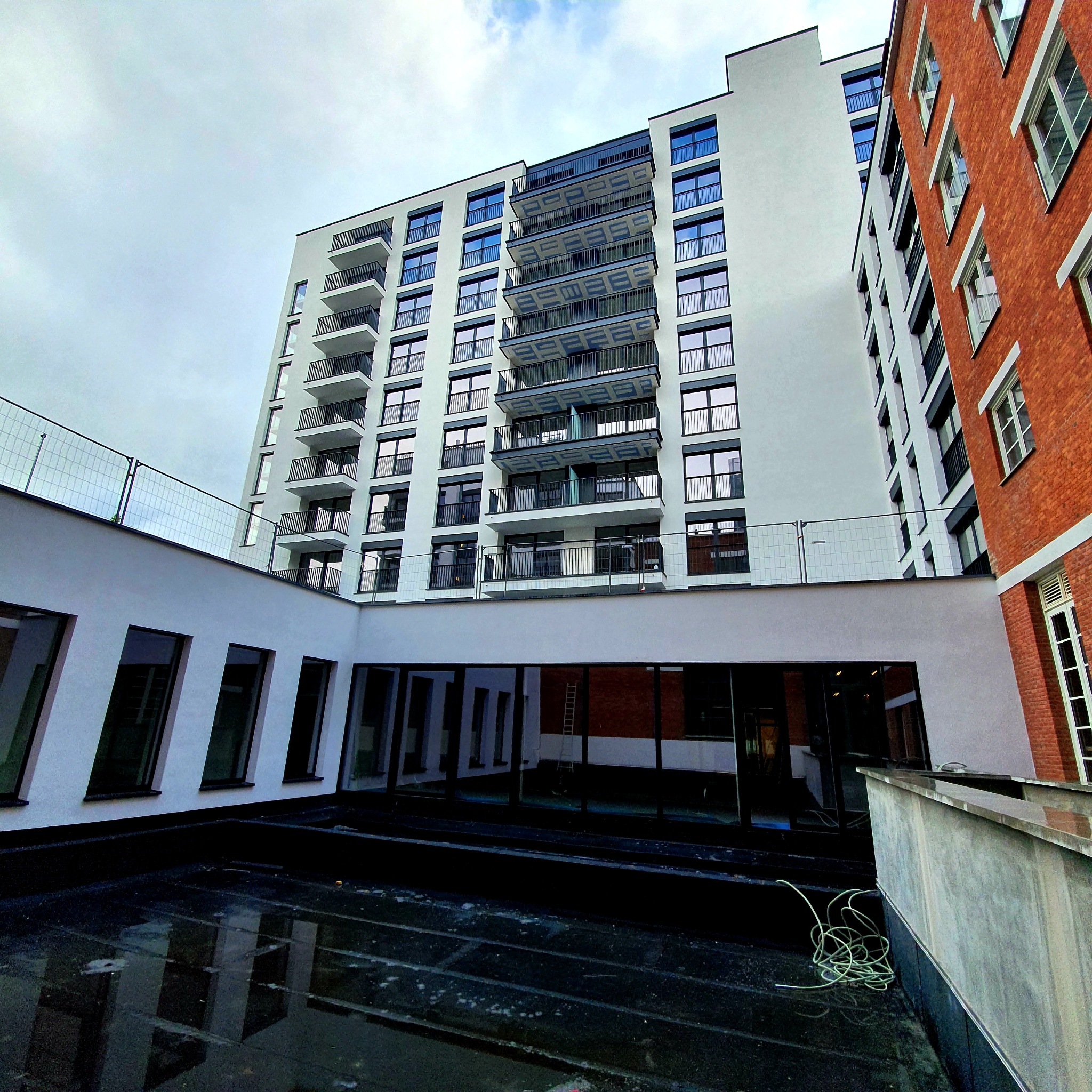 Wysoki budynek z białymi ścianami i balkonami, sąsiadujący z budynkiem z czerwonej cegły, widok z dołu na tle pochmurnego nieba, w dole widoczne odbicia w kałużach na dachu.