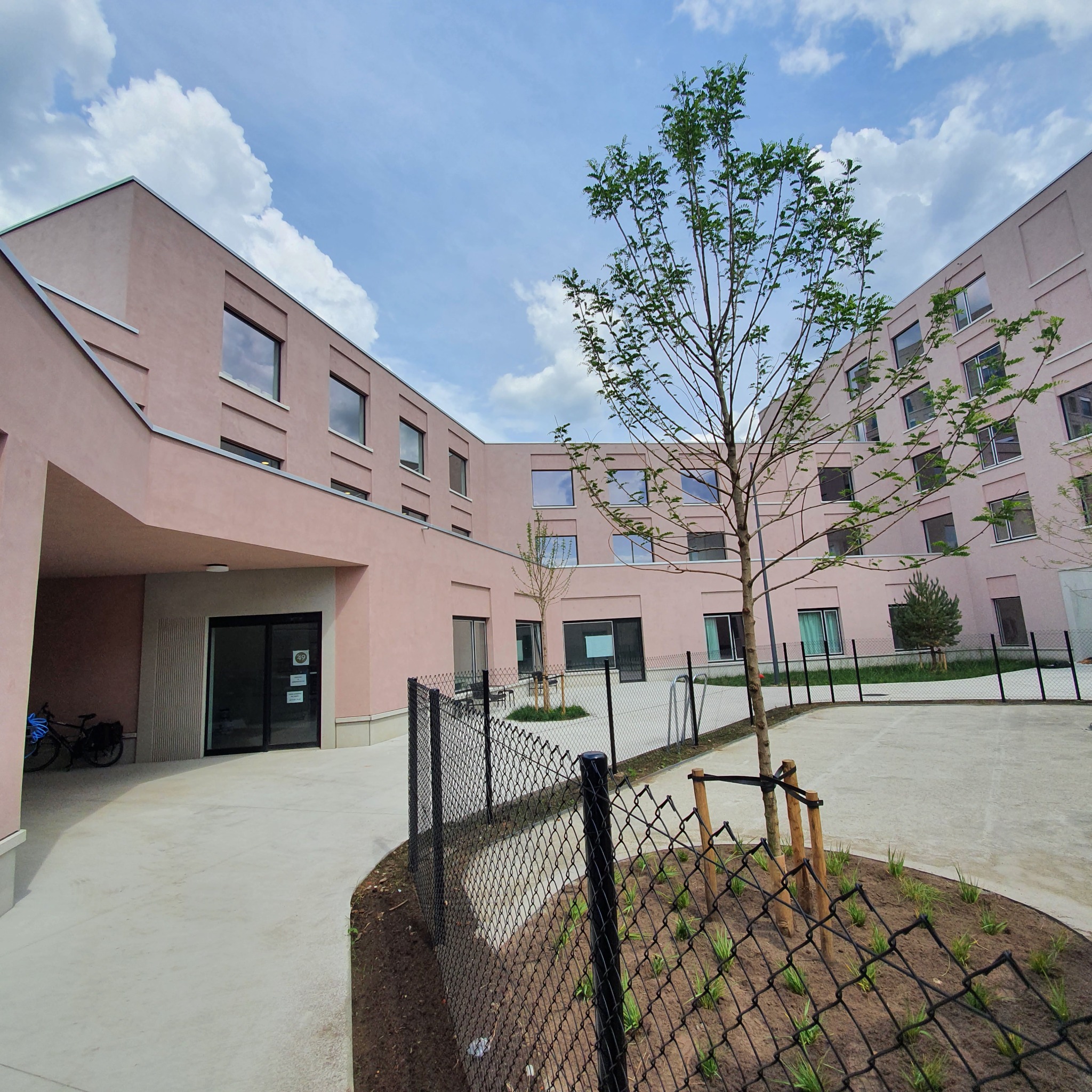 Nowoczesny budynek o różowej elewacji widziany z dziedzińca z młodymi drzewami, czarnym ogrodzeniem i błękitnym niebem z chmurami.