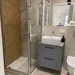 Nowoczesna łazienka z kabiną prysznicową z drewnianą ścianą, szarą szafką pod umywalką i widokiem na stół w jadalni w tle.