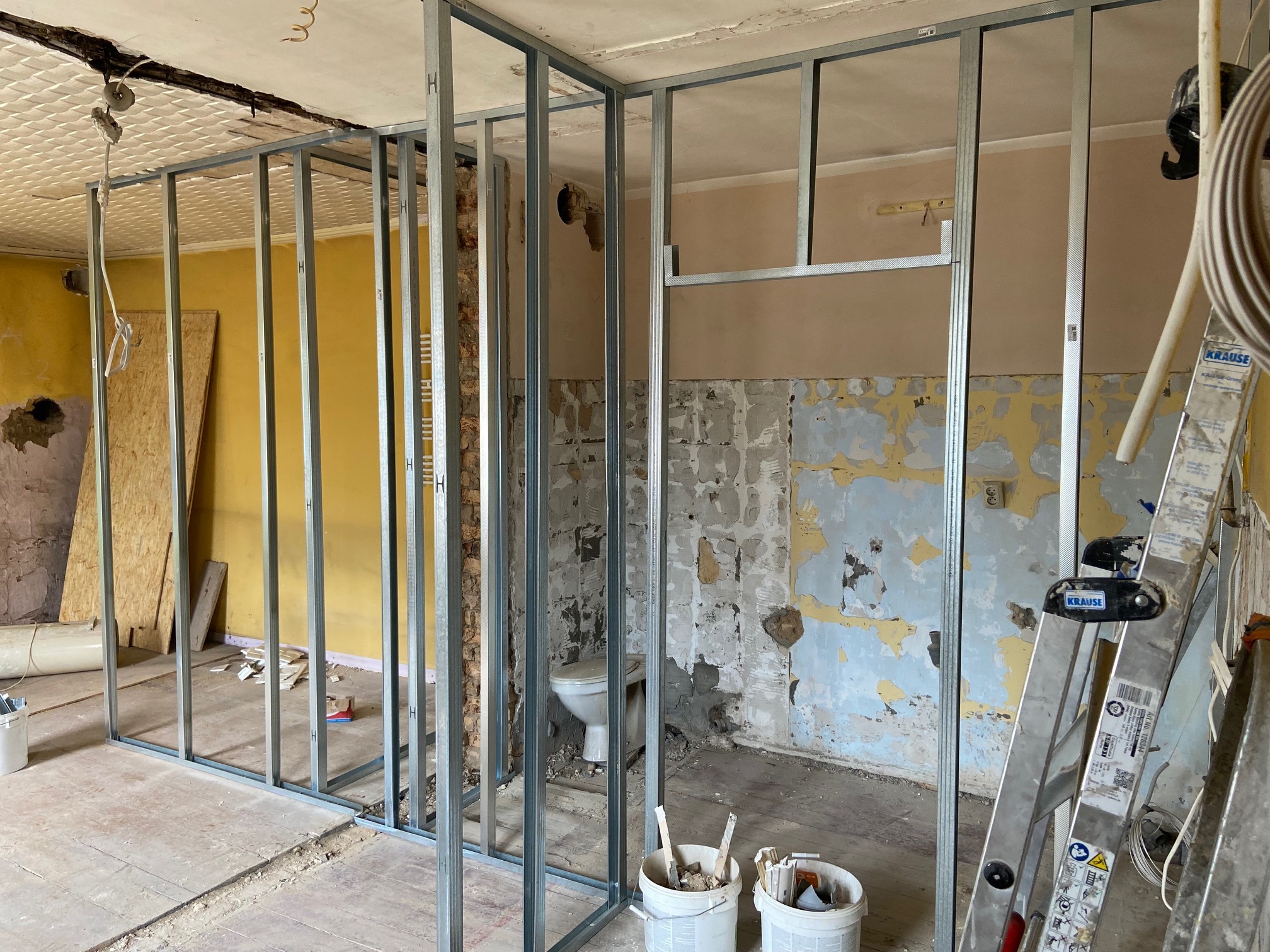 Stelaż z profili metalowych tworzący nową ścianę działową w trakcie remontu mieszkania, widoczne ślady starej farby na ścianach, drabina Krause i wiadro z narzędziami.