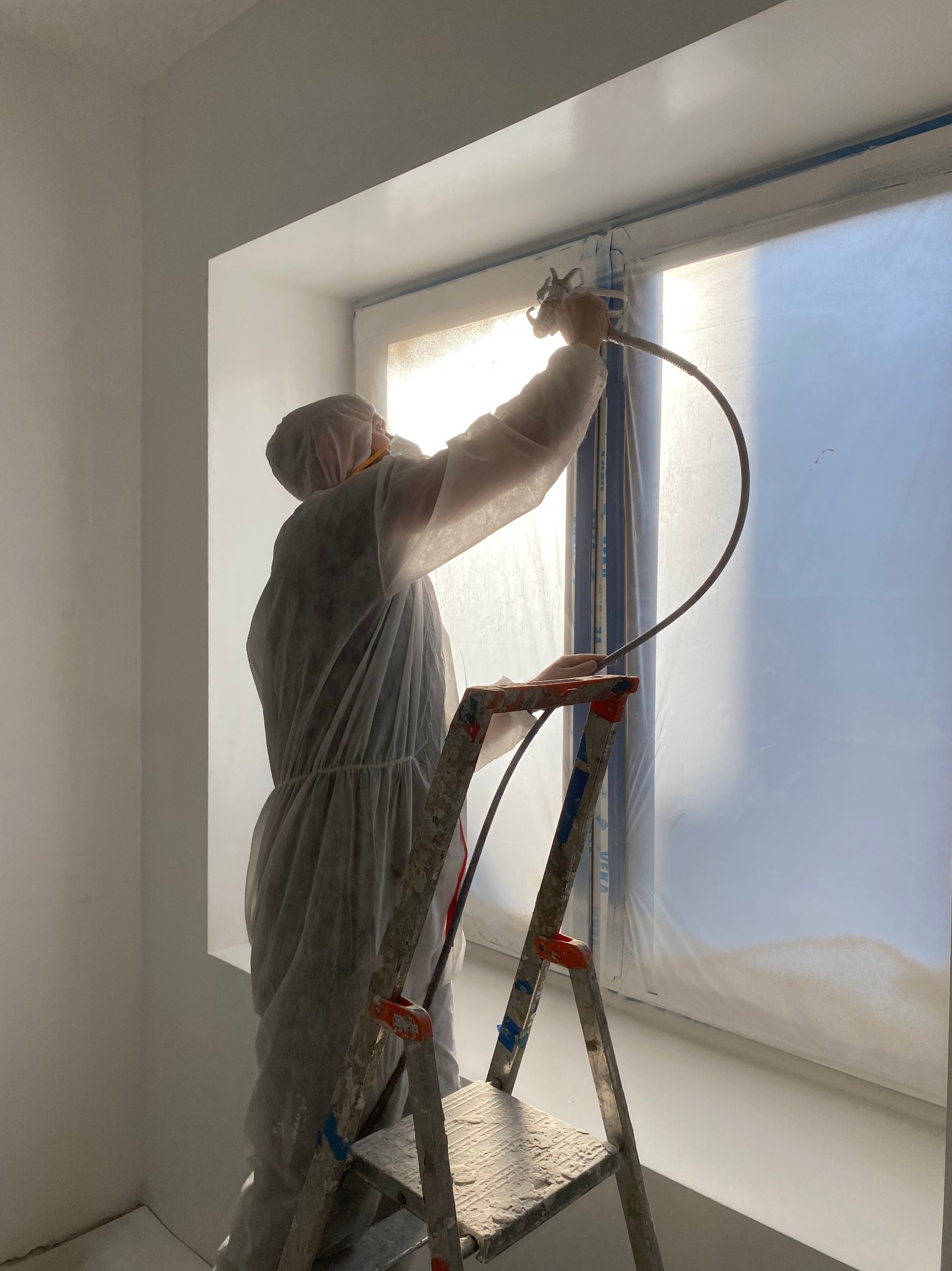 Pracownik w kombinezonie ochronnym maluje ramę okienną natryskowo, stojąc na zabrudzonej farbą drabinie w jasnym pomieszczeniu.