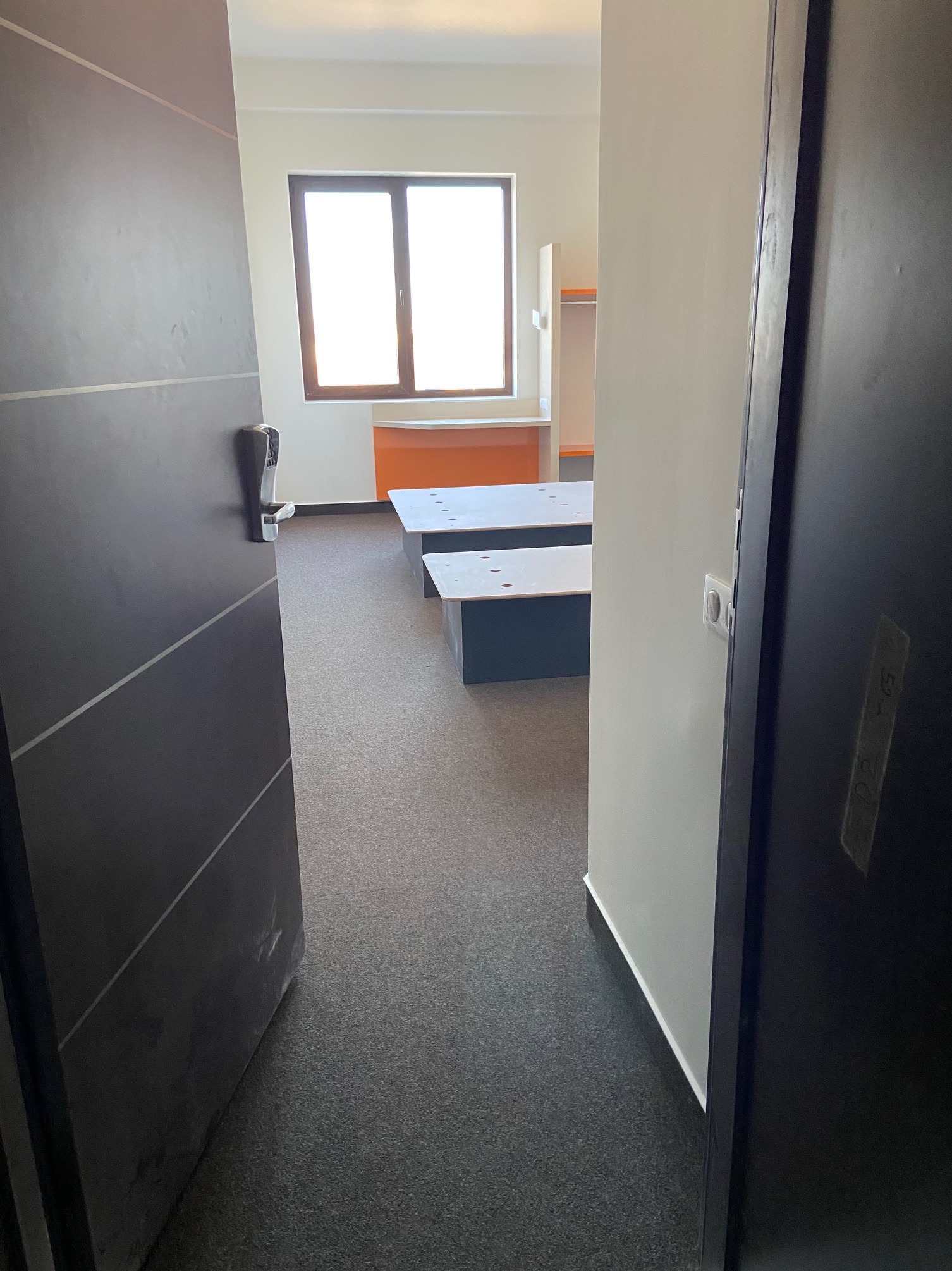 Widok zza uchylonych drzwi na nowocześnie wykończone wnętrze z szarą wykładziną, białymi ścianami i meblami w jasnych kolorach, widoczne ślady po malowaniu na drzwiach