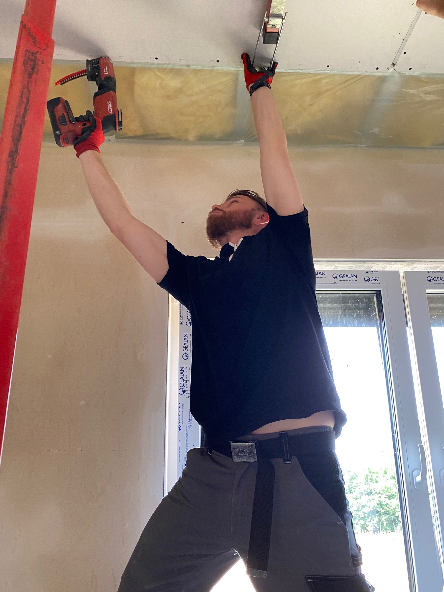 Pracownik w czerwonych rękawicach montuje płytę gipsową do sufitu za pomocą sztyfciarki Hilti, widoczna izolacja z wełny mineralnej.