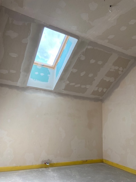 Pokój na poddaszu w trakcie szpachlowania, widoczne łączenia płyt gipsowo-kartonowych i okno dachowe z widokiem na błękitne niebo.