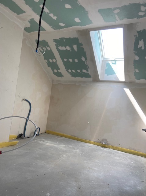 Poddasze z oknem dachowym w trakcie szpachlowania, widoczne łączenia płyt gipsowo-kartonowych wypełnione masą szpachlową, wystające przewody elektryczne.