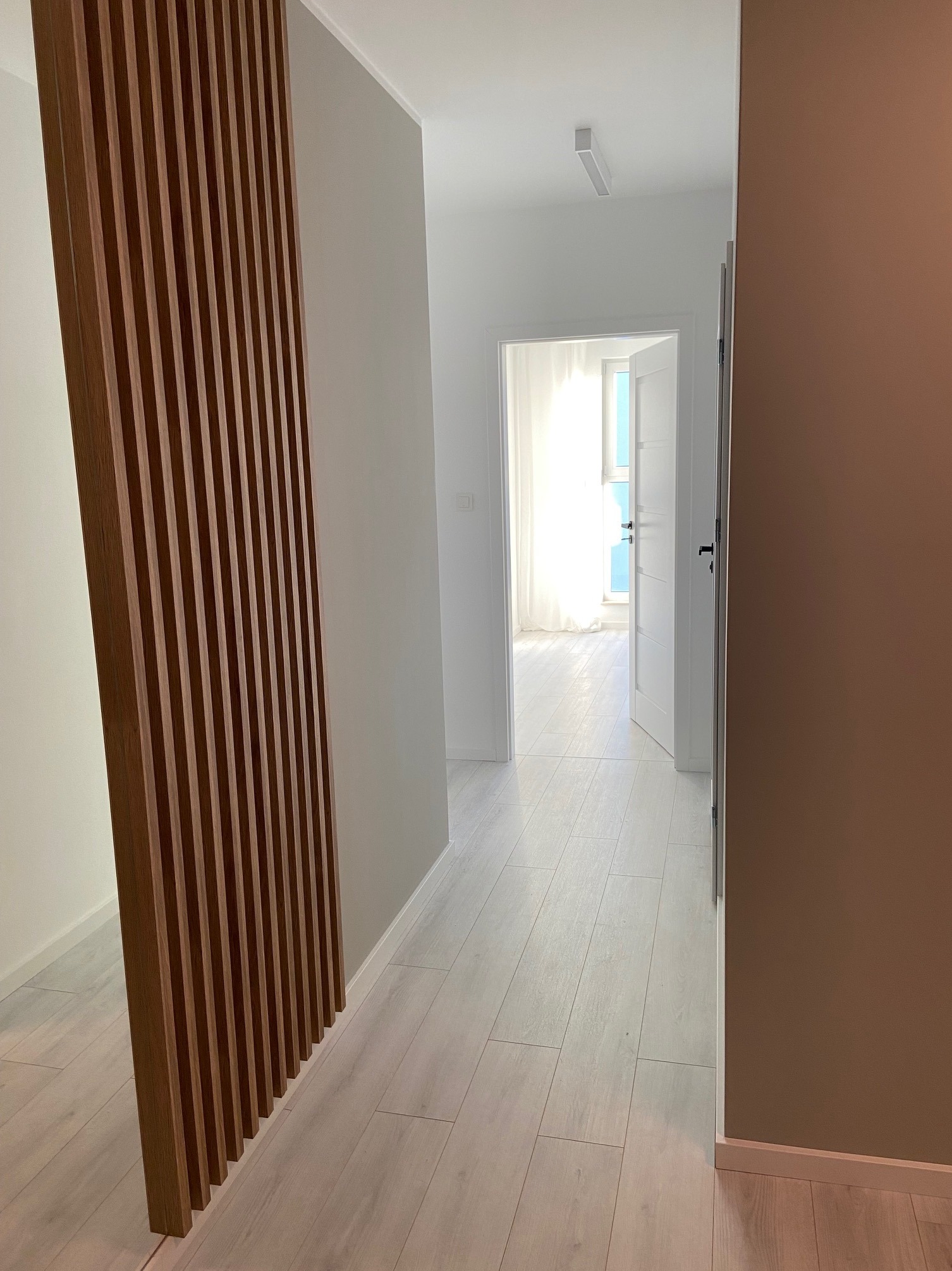 Jasne wnętrze mieszkania z drewnianą ażurową ścianką działową i świeżo pomalowaną ścianą w kolorze ciepłego brązu, widok na otwarte drzwi balkonowe w tle.