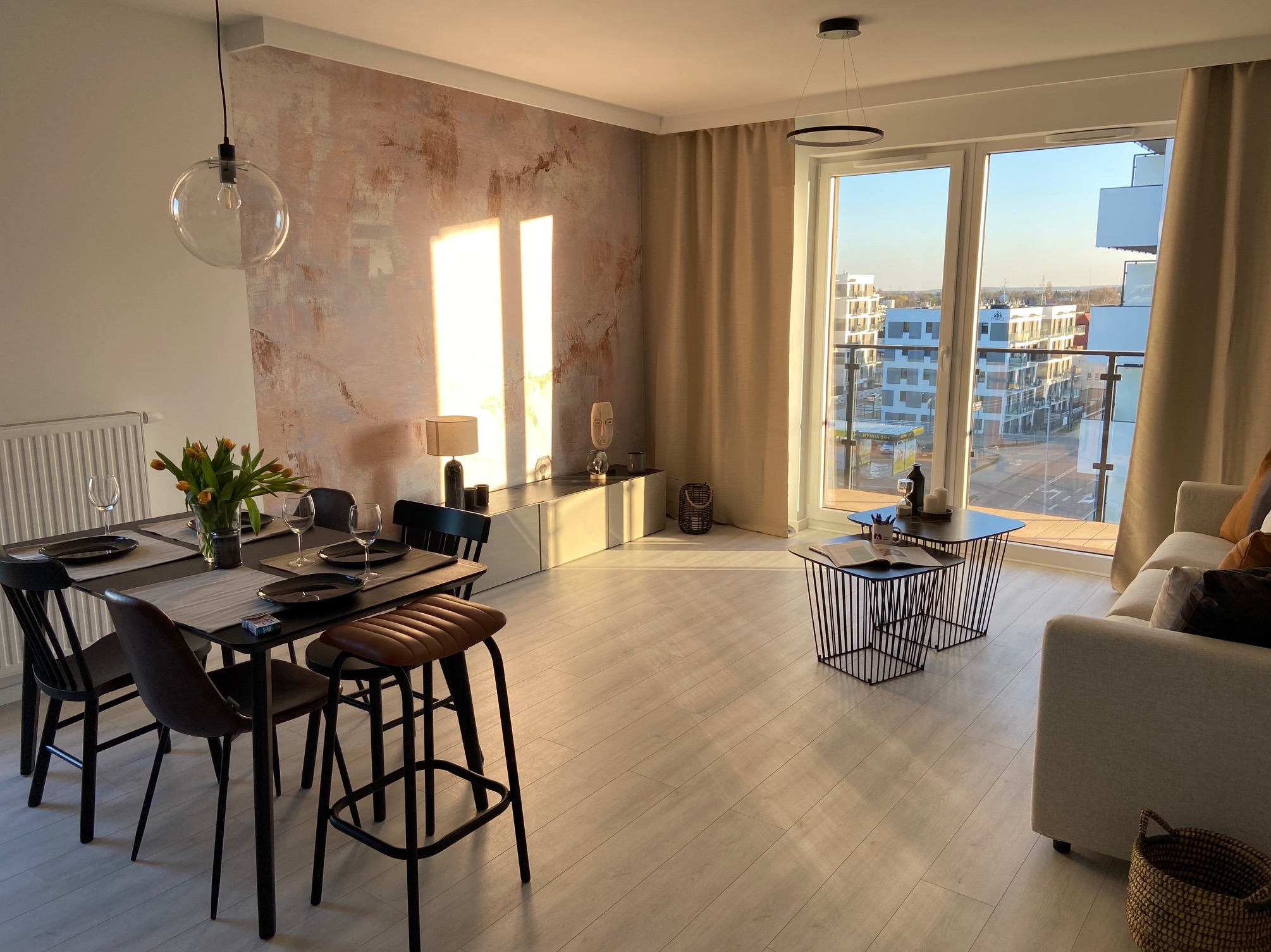 Jasne wnętrze mieszkania z dekoracyjną ścianą w odcieniach różu i brązu, nowoczesne meble, stół z nakryciem i bukietem tulipanów, widok na miasto przez duże okno.