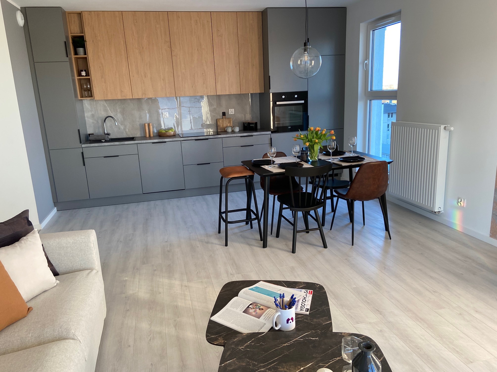 Nowoczesna kuchnia z szarymi szafkami i drewnianymi frontami, stół z czarnym blatem i tulipanami, widok na salon z sofą i stolikiem kawowym z książkami.