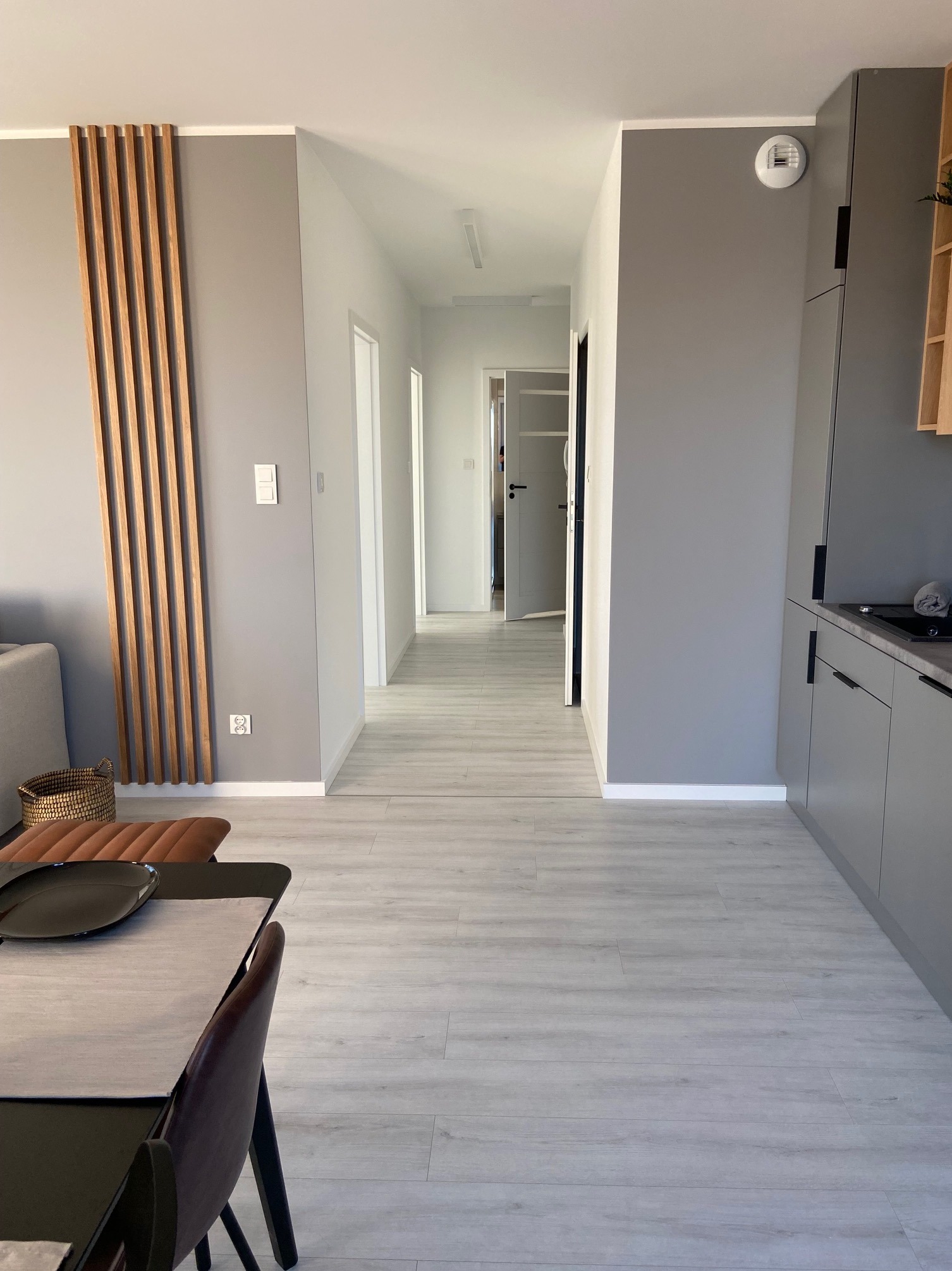 Nowoczesne wnętrze mieszkania po remoncie: szare ściany, jasna podłoga imitująca drewno, dekoracyjne drewniane listwy na ścianie, widok na korytarz z otwartymi drzwiami.