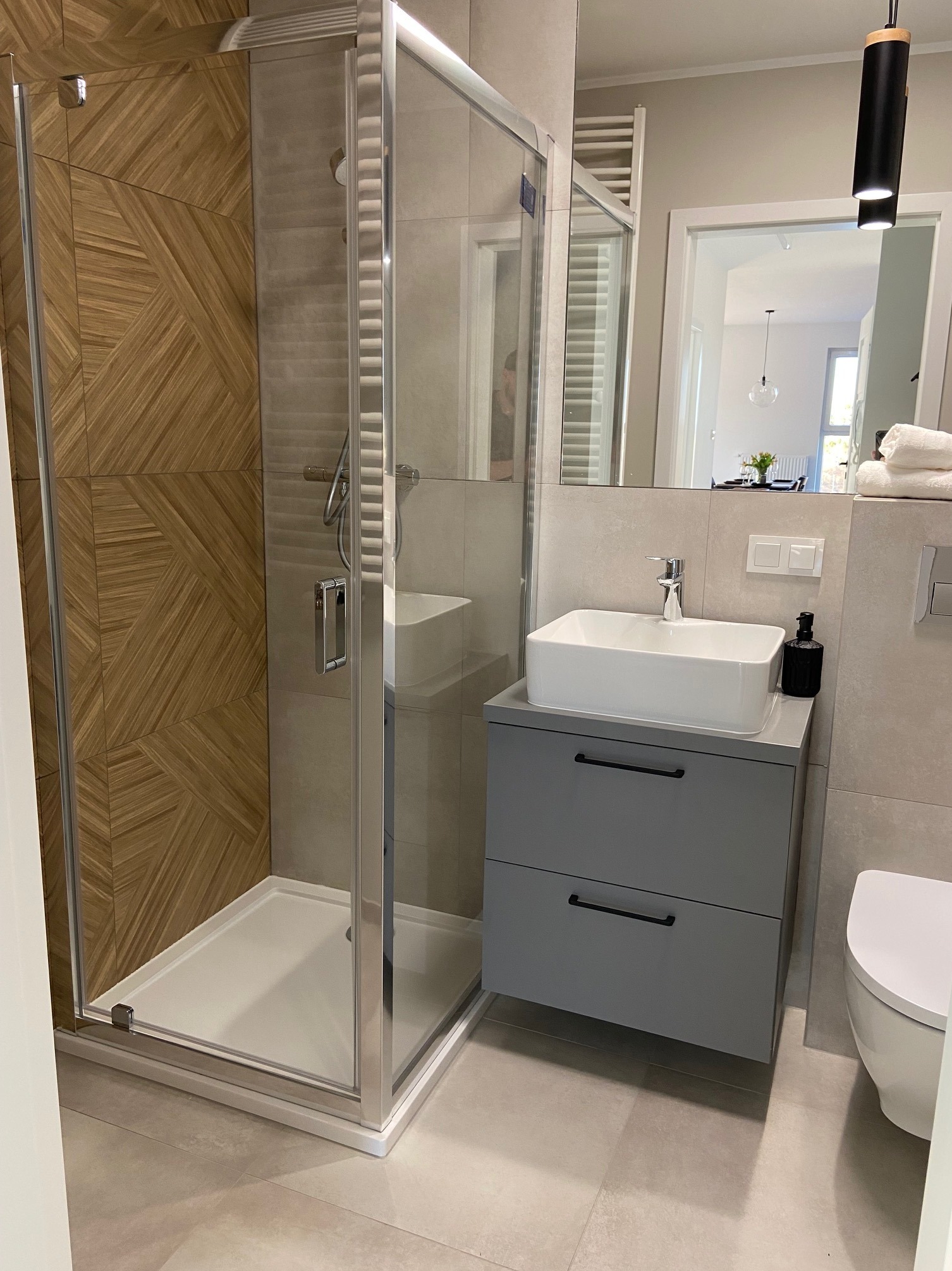 Nowoczesna łazienka z kabiną prysznicową z drewnianą ścianą, szarą szafką pod umywalką i widokiem na stół w jadalni w tle.