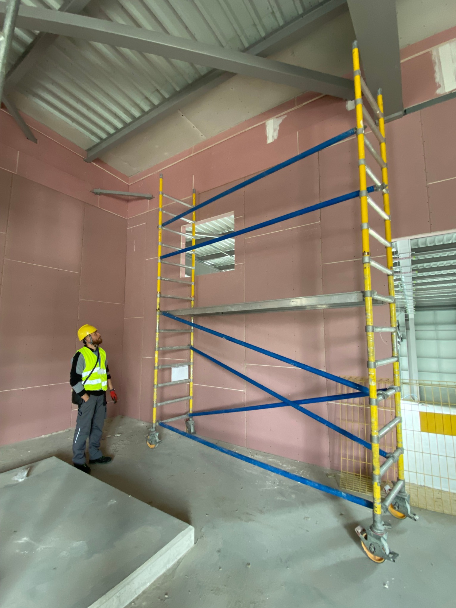 Pracownik w kasku i kamizelce odblaskowej stoi obok rusztowania na tle ściany przygotowanej do malowania, z widocznymi płytami gipsowo-kartonowymi.
