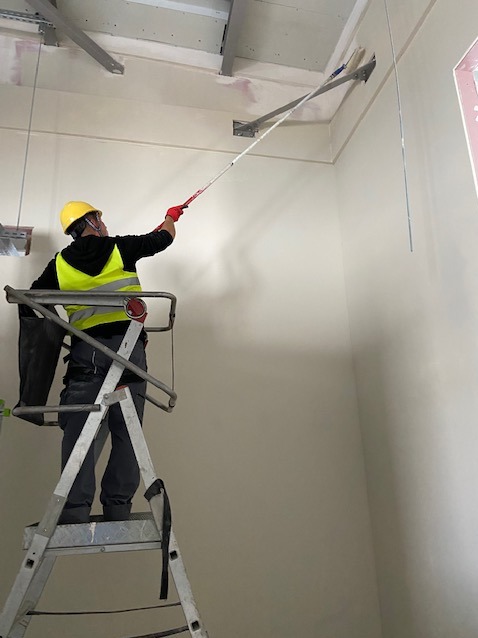 Pracownik w żółtym kasku i kamizelce odblaskowej maluje sufit w pomieszczeniu za pomocą wałka na długim wysięgniku, stojąc na aluminiowej drabinie.