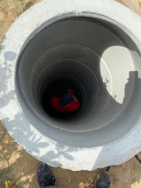 Wnętrze betonowej studni z pracownikiem w czerwonej koszuli i niebieskiej czapce na dnie, widok z góry.