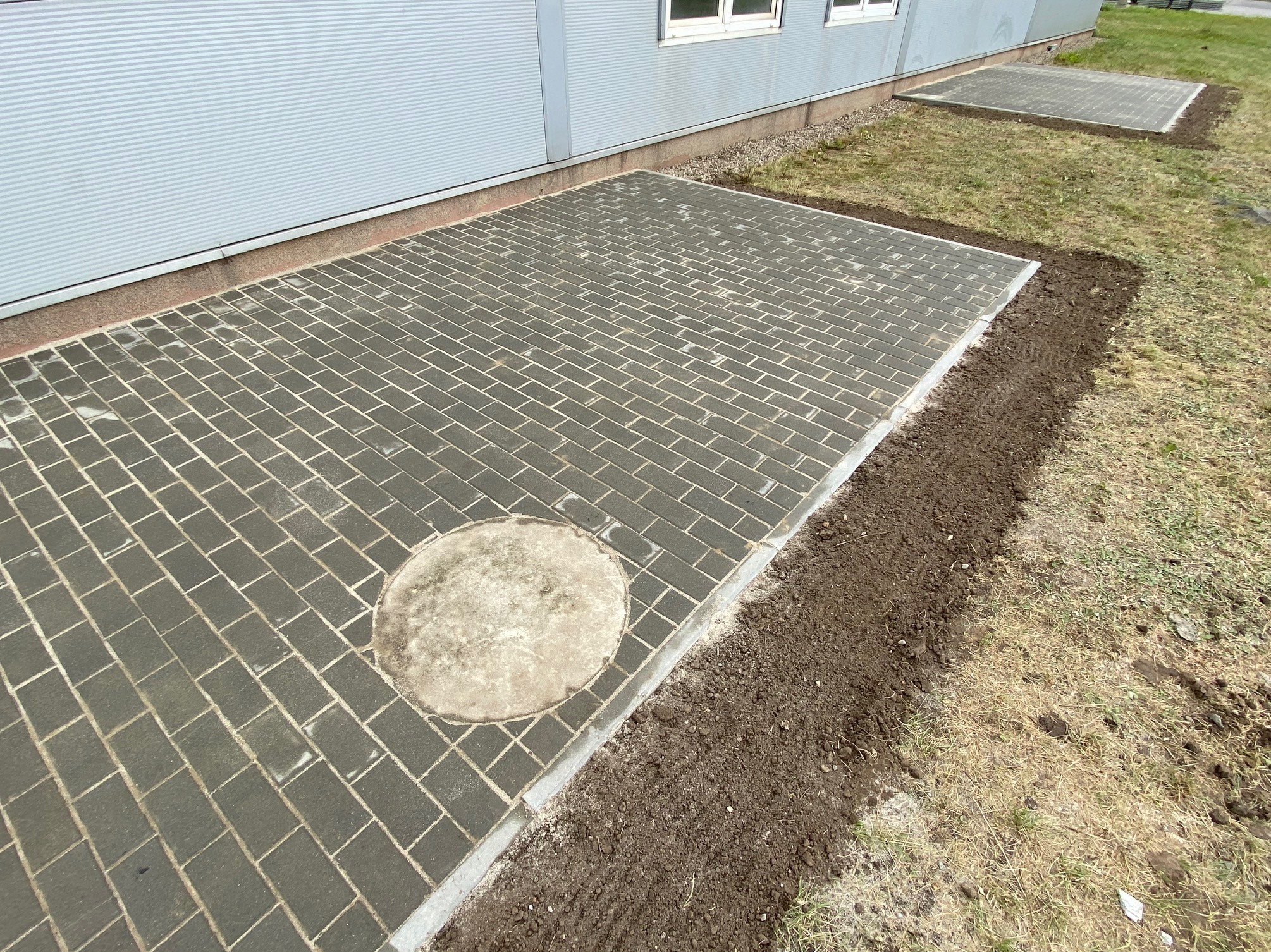 Ułożona szara kostka brukowa na podwórku z okrągłą studzienką w rogu, obok budynek z blachy falistej.