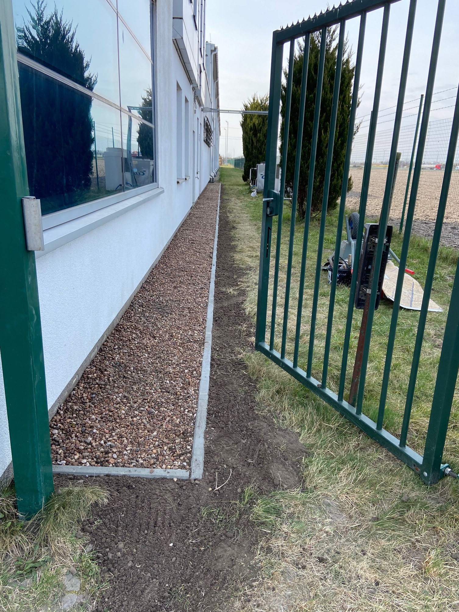 Wykończony fragment terenu przed budynkiem z białym tynkiem, wysypany drobnymi kamieniami i obłożony betonową obwódką, widok zza zielonej, otwartej bramy.