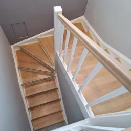 Kątowe ujęcie schodów drewnianych z białymi balustradami, widoczne świeżo wycyklinowane stopnie zabezpieczone taśmą malarską.