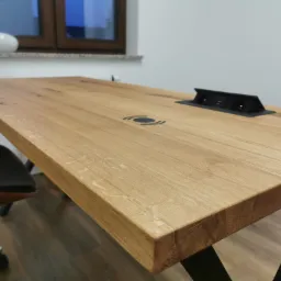 Jasny, dębowy blat biurka z czarnymi, metalowymi nogami w kształcie litery X, wbudowanym gniazdkiem elektrycznym i ładowarką indukcyjną, widok z perspektywy