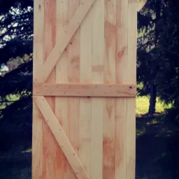 Surowe, drewniane drzwi z ukośnymi wzmocnieniami, wykonane z jasnych desek, umieszczone na zewnątrz na tle zieleni.