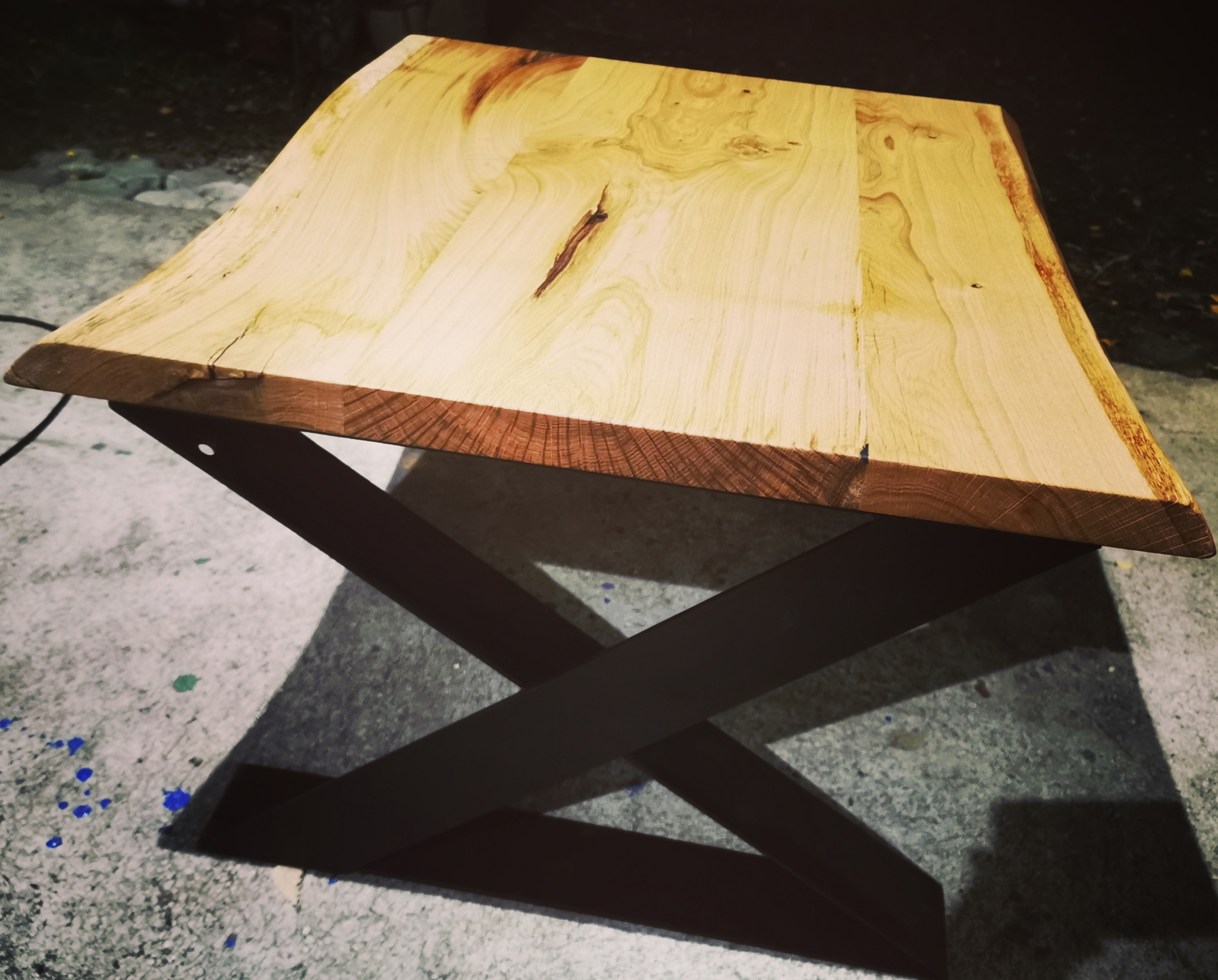 Niski stolik kawowy z blatem z jasnego drewna o nieregularnych krawędziach i nogami w kształcie litery X, rzucający cień na podłogę z plamami farby.