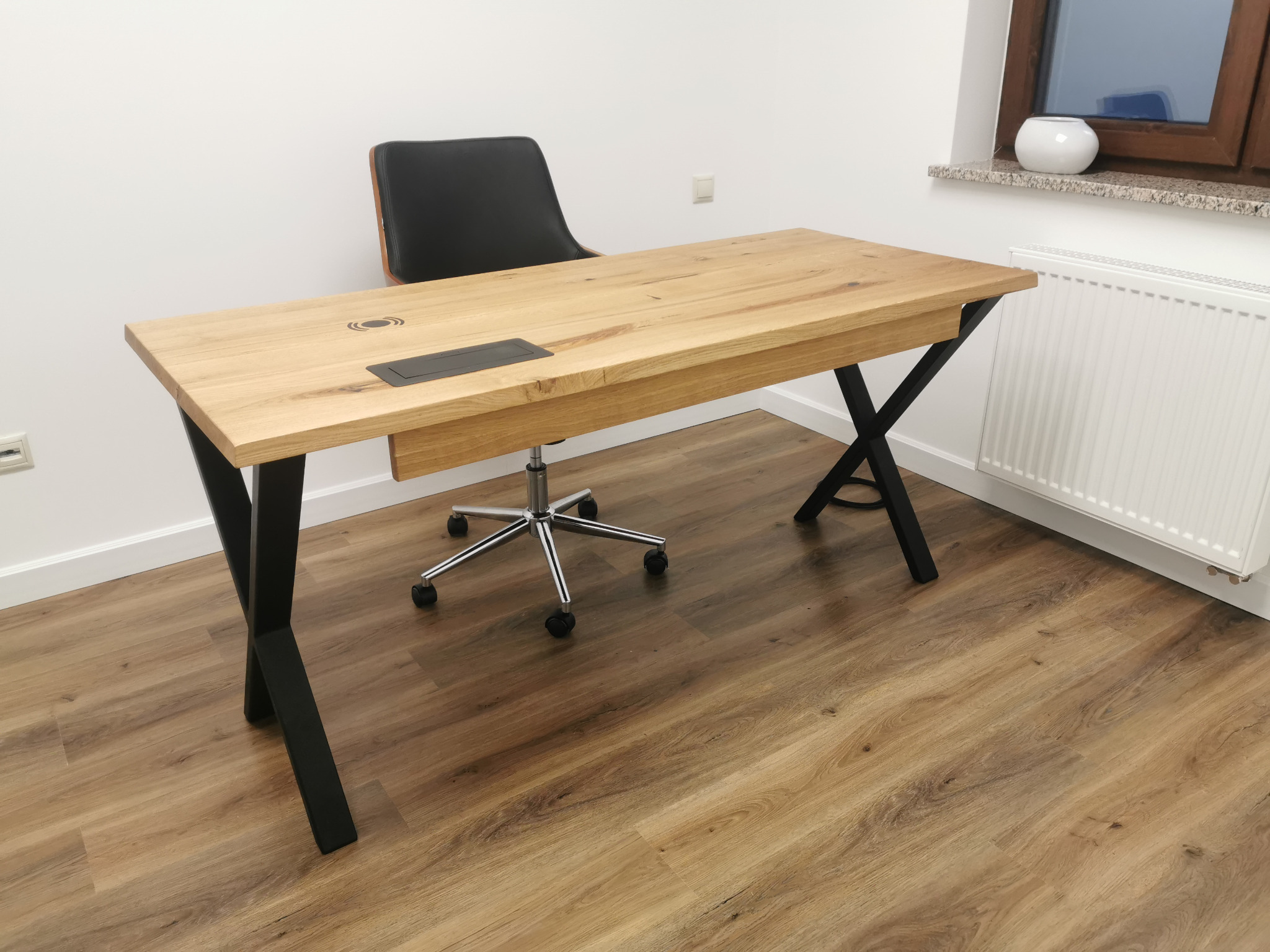 Biurko z blatem z jasnego drewna i czarnymi nogami w kształcie litery X, z wbudowanym gniazdem i ładowarką indukcyjną, na podłodze z paneli, w tle krzesło biurowe i grzejnik.