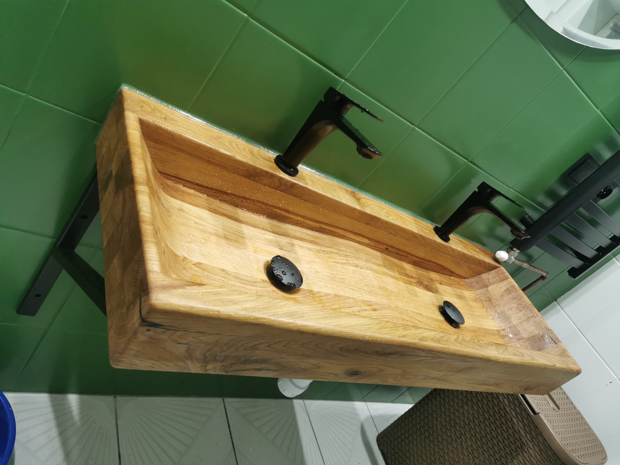 Drewniana umywalka nablatowa z czarną armaturą, zamontowana na metalowym stelażu, na tle zielonych płytek ściennych.