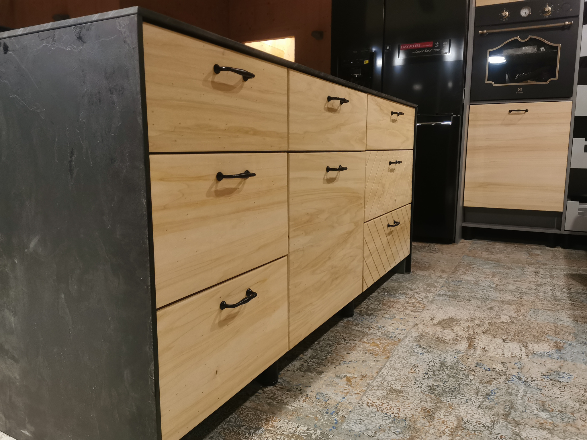 Blat kuchenny z szufladami wykonanymi z jasnego drewna, z czarnymi uchwytami, wykończony czarnym blatem o teksturze kamienia, na tle czarnej lodówki i piekarnika w stylu retro.