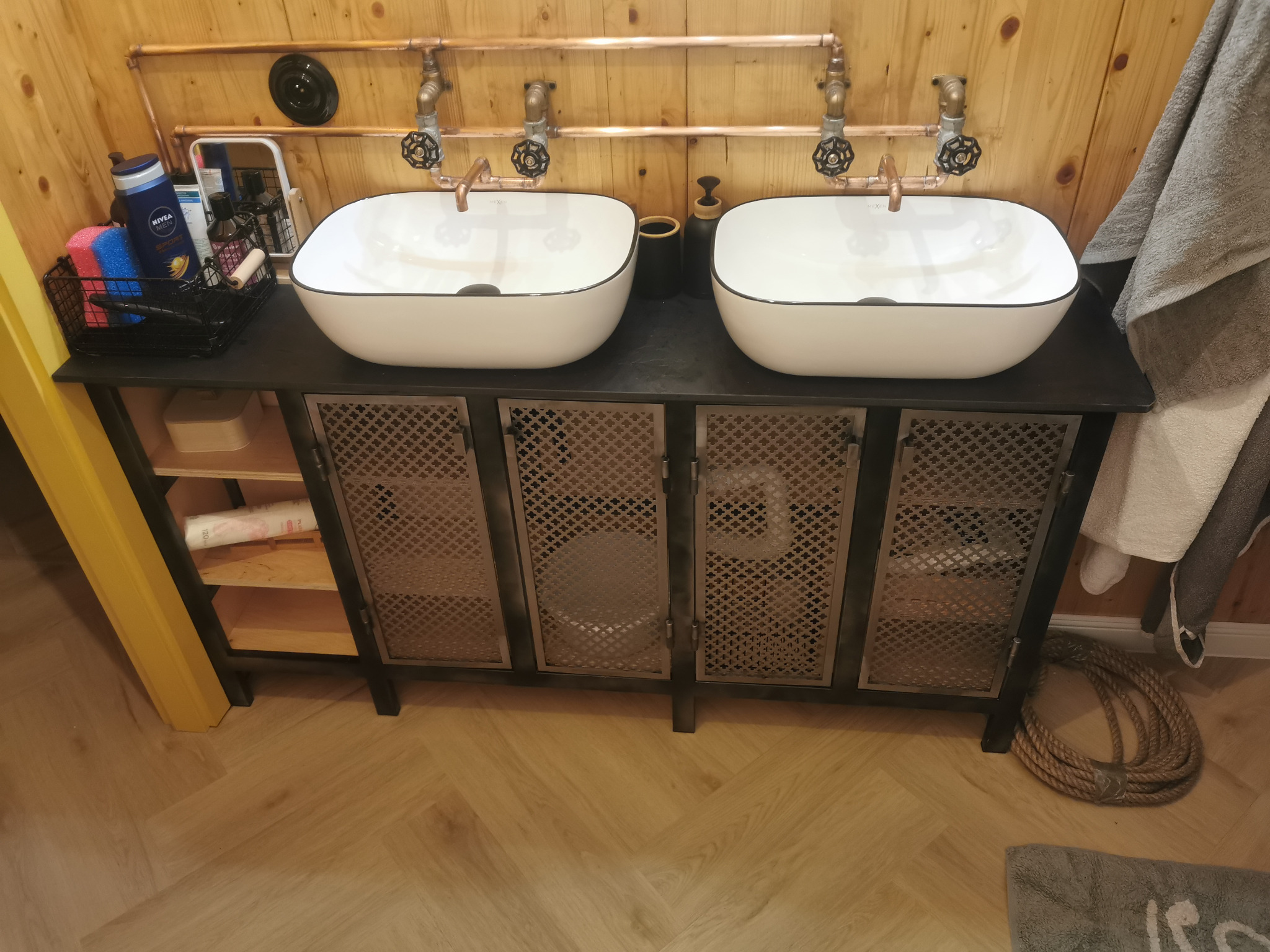 Łazienka w stylu rustykalnym z dwoma umywalkami nablatowymi w białych misach z czarnym obramowaniem, armaturą z miedzianych rur, szafką z metalowymi drzwiczkami z ażurowym wzorem oraz drewnianymi...