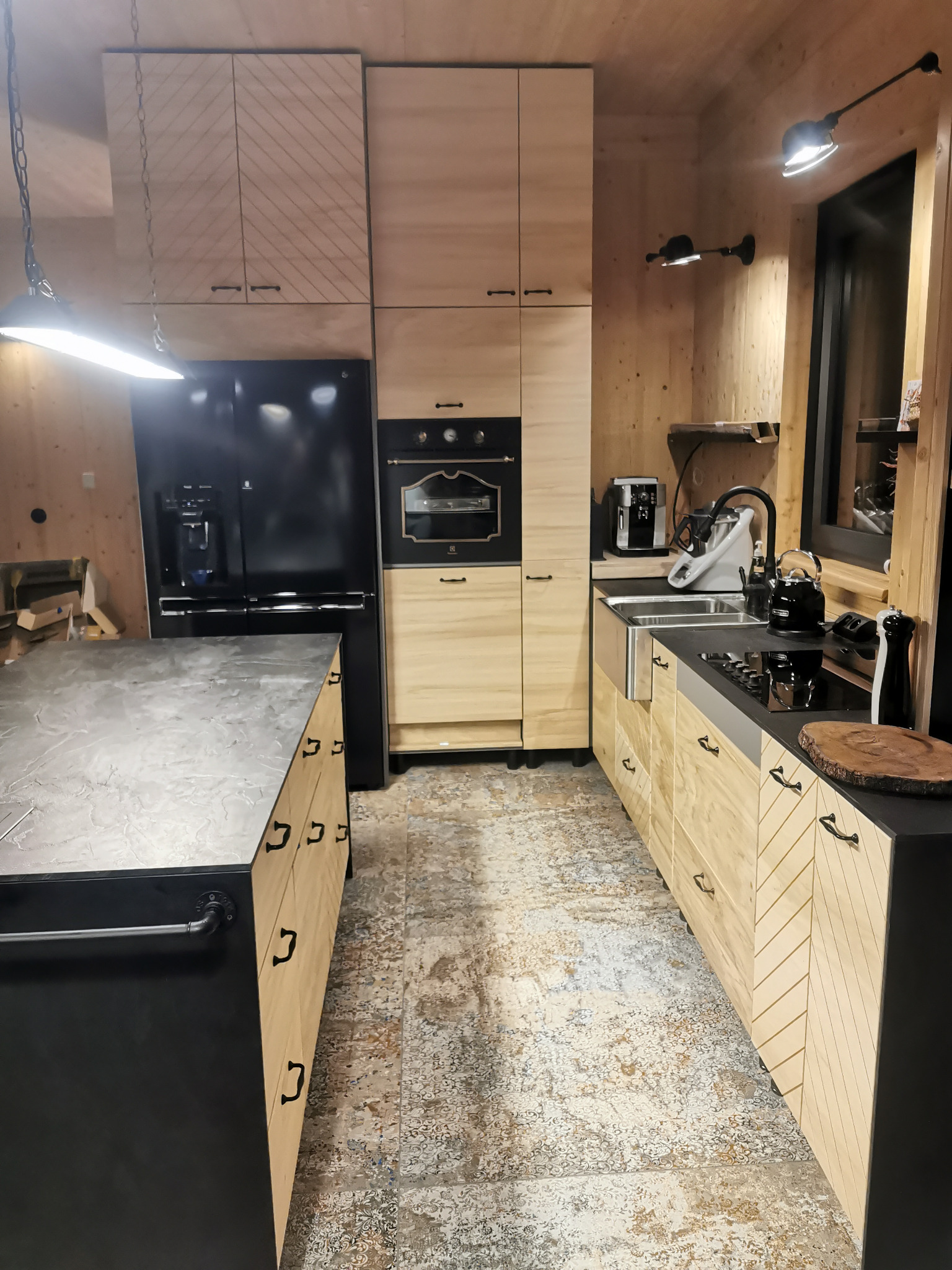 Wnętrze kuchni z drewnianymi szafkami o jasnym odcieniu i wzorze w jodełkę, czarnym blatem, wyspą kuchenną z szarym blatem, czarną lodówką i oświetleniem w stylu industrialnym.