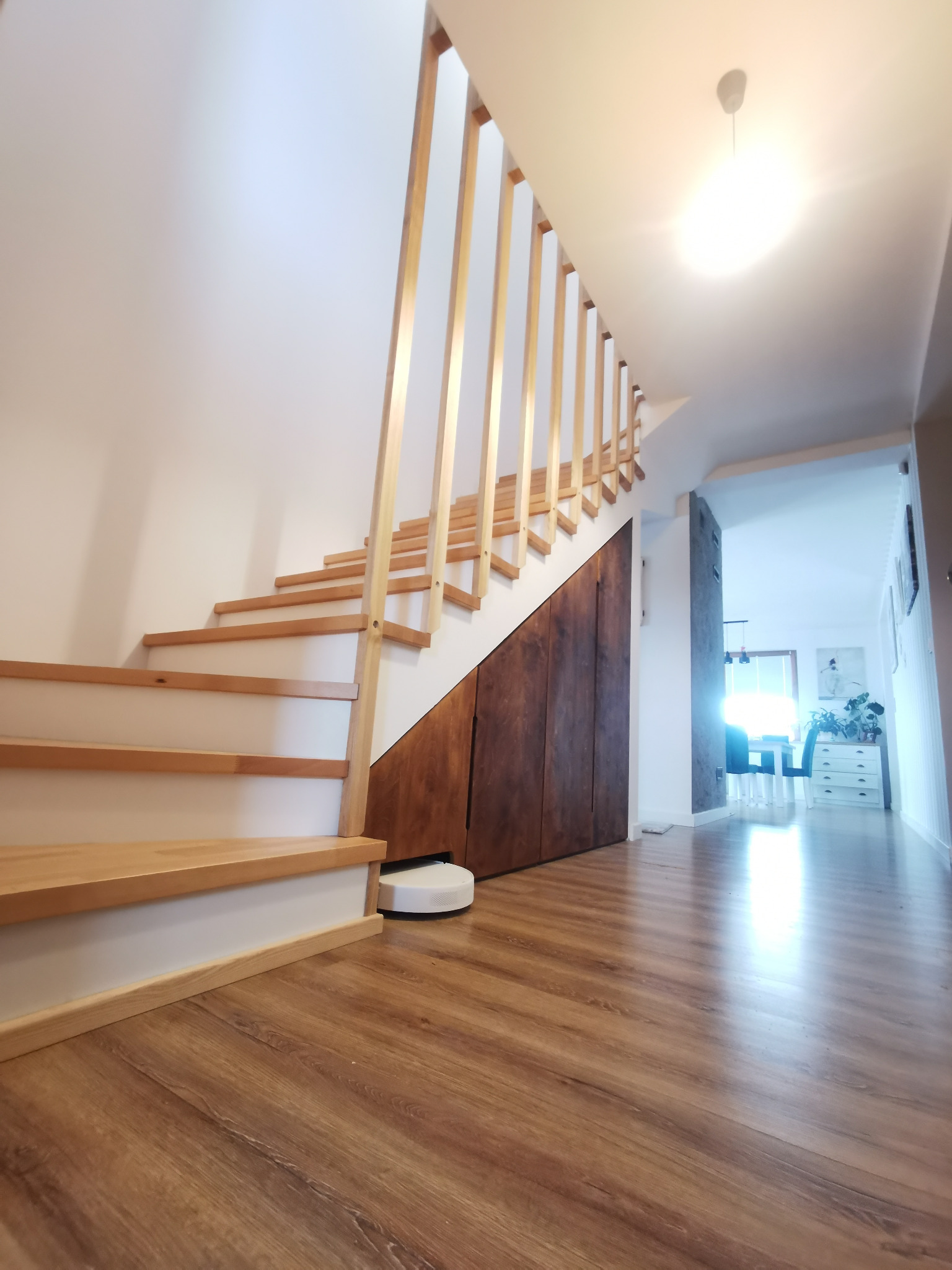 Drewniane schody z jasnymi stopniami i balustradą w Ostrołęce, zintegrowane z ciemną szafą wnękową pod schodami, na podłodze robot sprzątający.